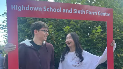 Highdown School and Sixth Form Centre A boy and girl smile at one another. They are holding a red rectangle that is framing both of them in shot. At the top of the rectangle the words "Highdown School and Sixth Form Centre" are printed. 