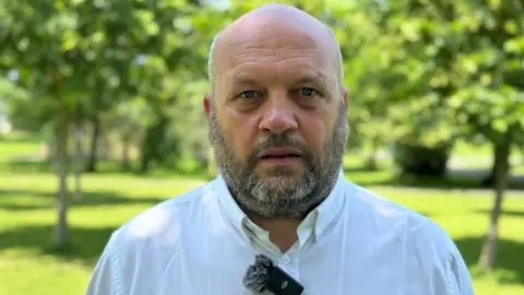 Barry Taylor, a man with a shaved head and a grey beard, looks at the camera while standing in a park on a bright sunny day. He is wearing a white open-necked shirt.