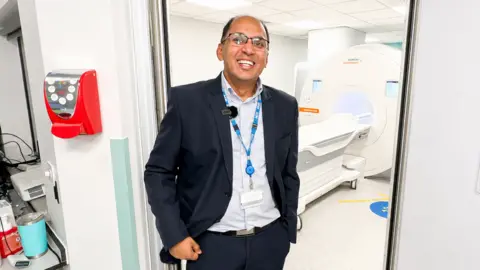 Robin Joseph is wearing glasses and has a suit on, with an MRI scanner behind him.