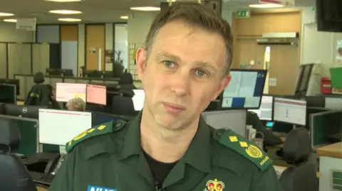 Adrian South is wearing a green ambulance uniform and is looking directly at the camera. There are computer monitors around him and desks with people sat at them. 