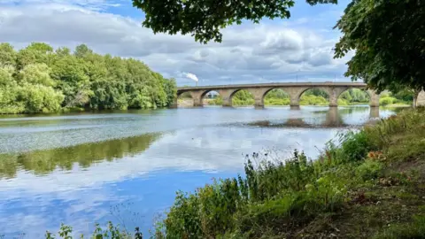 Wander Vision A broad, flat river, with a grassy bank on one side, and trees - in full leaf - reaching down to the water on the other. It is spanned by a stone bridge with seven arches. The sky, blue in patches with high clouds, is reflected in the river.