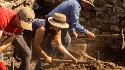 Two men and a woman digging in the sunshine at the Auckland Palace excavation site. They are wearing hats and leaning as dust flies in the air.
