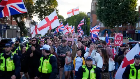 PA Protesters marching in a tight group, marshalled by a line of police officers at the front. The protesters are waving England and Union flags. There are a handful of women and children near the front of the crowd.