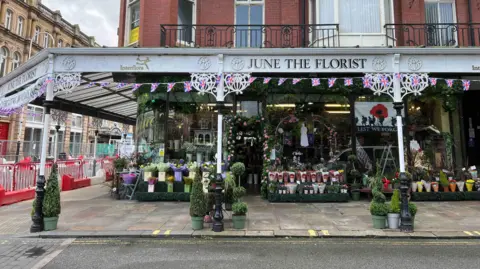 The outside of the shop, it has a white canopy with June the Florist written in gold lettering. The front is covered in flowers.