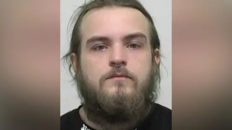 Mugshot of Kennedy. He has a straggly beard and long messy brown hair, with a ring through the septum of his nose.