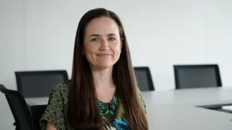 Bethan Evans with long dark hair is sitting in an office with white walls, a desk and black chairs in the background.