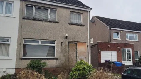 A rundown home, with the front door boarded up, and overgrown greenery across the front