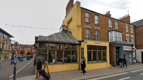 A screenshot from Google Street View showing a yellow restaurant building with large windows on a street corner, with a few people walking on the pavement in front of it. To the left of it is a road sign banning cars and motorcycles from turning into the road.