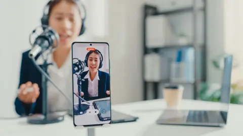 Getty Images A person speaks into a microphone while being recorded on video by a mobile phone, while sitting at a white desk in a room with a laptop open and coffee cup on the table, and a bookcase in the background.