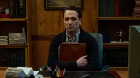 Apple TV Matthew Rhys in Widow's Bay. He is sat down, holding a folder, looking straight ahead 