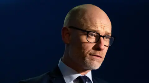 Matthew Horwood/BBC A headshot image of a man wearing a shirt with a tie and a dark suit jacket over it. He is wearing glasses and looking past the camera. 