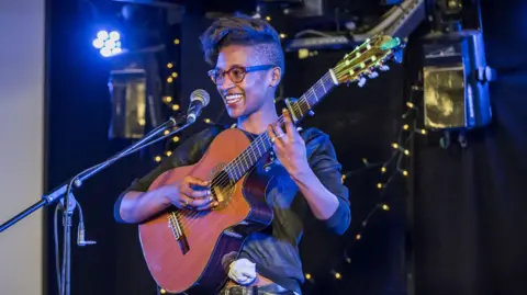 Chris Scott, Flint & Pitch A musician stands on a small stage holding an acoustic guitar beside a microphone, illuminated by blue stage lighting. The dark venue background is decorated with warm string lights, creating an intimate live-performance setting.