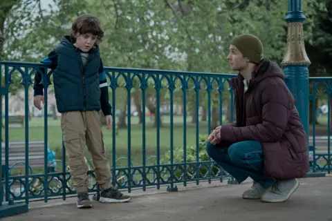 Duck Soup Films A young boy is leaning on a railing, as a man is bent down trying to speak with him.