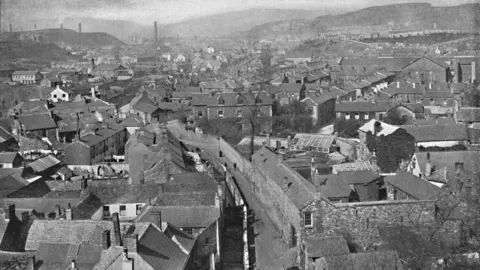 Getty Images Merthyr Tydfil, circa 1896, it is a view of the town in black and white with densely packed terraced housing and industrial chimneys in the distance. Hills can be seen in the distance.