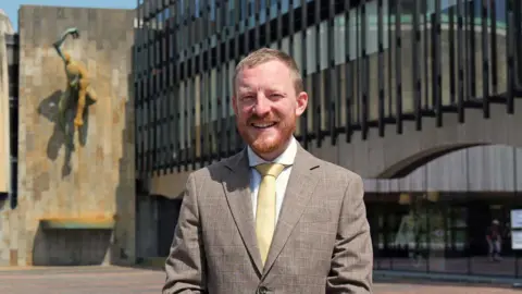 LDRS Newcastle City Council's Liberal Democrat leader Colin Ferguson, a man with fair hair and a beard, is smiling at the camera. He is wearing a light coloured suit jacket, white shirt and pale yellow tie.