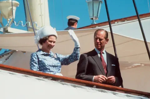 Getty Images A man and a woman stand together on a boat. The women is wearing a blue dress, a white hat and white gloves. She is waving her hand. The man is in a black suit with a red tie.