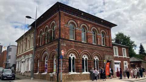 A large two storey red brick building, with arched window frames. There are people stood outside the main entrance