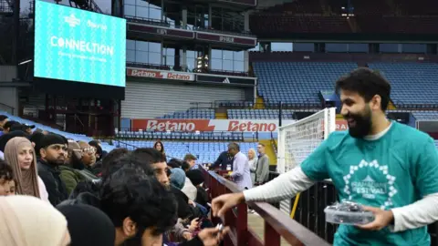 Huzayfa Dawood A man wearing a turquoise t-shirt hands out dates to people sat in a football stand.