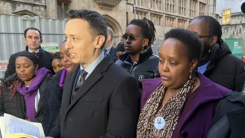 DCI Mark Almond delivering a statement to press outside court, reading from an open notebook in his hands. The family of Eddie Kinuthia, including his mum Irene, are surrounding him and look very solemn. They are all wearing a purple item of clothing, Eddie's favourite colour.