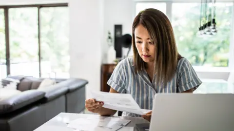Woman wearing striped shirt looking at bills while sitting at laptop in her home