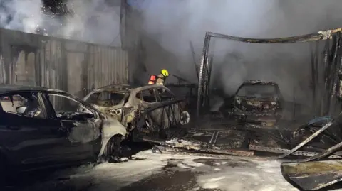 South Yorkshire Fire & Rescue A firefighter can be seen among the burnt out shells of three vehicles. It is dark and there are large pools of water on the ground.