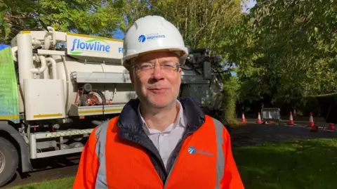 Vikki Irwin/BBC Paul West is wearing a high visibility orange jacket and is standing in front of a truck which is on a green. You can also see traffic cones and barriers in the background. Paul is wearing a hard hat which says Suffolk Highways on it. 