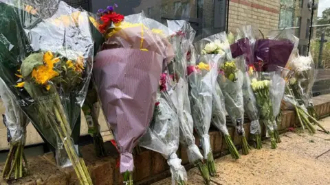 Phil Shepka/BBC A close-up of a row of bunches of flowers that have been placed on the ground in a street. The individual bunches are wrapped in plastic or paper. 