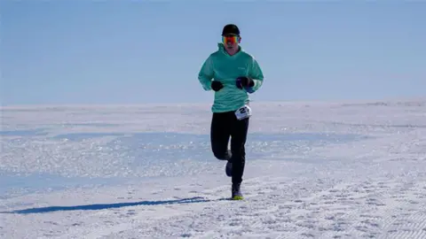 Mr Holborn, wearing an orange sun visor, teal hoodie and black trousers, running along the ice of Antarctica