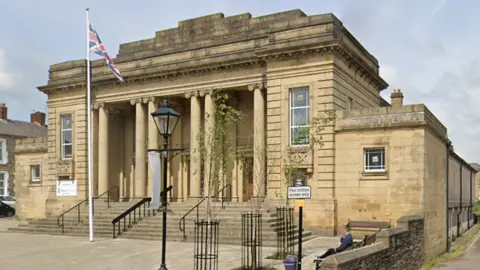 Mercer Hall is a large stone-built property with six large pillars at the entrance, above steps with black railings. It has a flagged frontage with a Union Jack flag flying from a flagpole and a vintage street lamp.