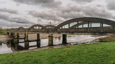 Getty Images An old arched bridge structure with water flowing underneath and grassy banks under an cloudy grey sky
