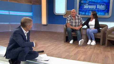 Jeremy Kyle sitting on a step on the set of a TV studio looking up at Steve Dymond sat in a brown tub chair. There is a Jeremy Kyle blue sign in the background.