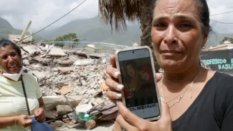 Woman crying shows picture of two missing daughters in front of destroyed building where they were last seen