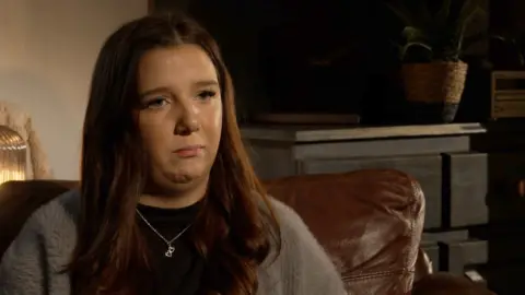 Ceri Owen is sitting on a brown leather chair. She has long brown hair, blue eyes and is wearing a silver necklace with two hearts