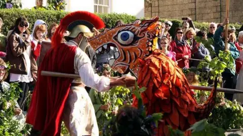 Helston Flora Day Association An actor dressed as a Roman with a helmet and cloak is jousting an actor wearing a dragon costume with a paper mache head. The majority of the people in the crowd surrounding them are smiling.