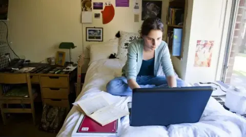 Getty Images Young woman sat cross-legged on a bed using a laptop with an open book in front of her to the right