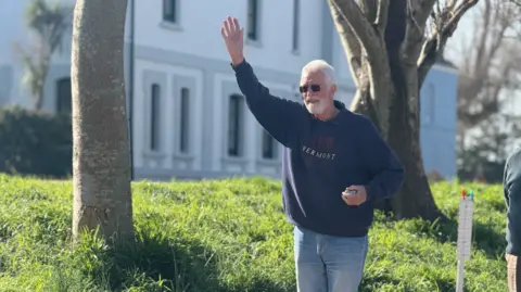 Rod Amy, in his 60s, outdoors on a sunny day, raising throwing a pétanque ball and has one arm in the air while holding another ball in the other hand. He's wearing a dark sweatshirt and jeans, standing beside a grassy area with trees and a white building in the background.