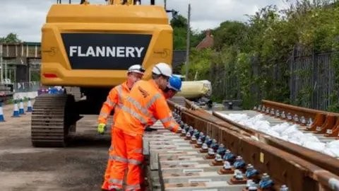 Network Rail employees dressed in orange overalls, wearing white and blue hats inspect new tracks before being lifted into position by a digger. 
