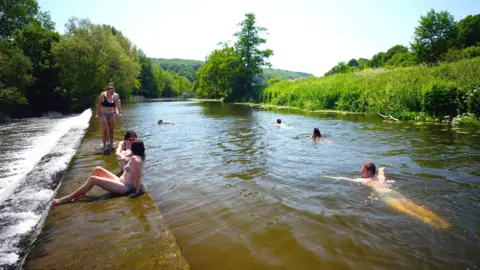 PA Media A small group of wild swimmers in the River Avon at Warleigh Weir. Four people are swimming in the river while three women are standing or sitting at the edge of the weir.