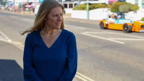 OPCC Jones by the side of a road wearing a blue dress. A sports car is in the distance.
