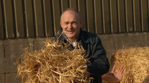 Martin Giles/BBC Sir Ed Davey, wearing a dark jacket, clutches a pile of hay.