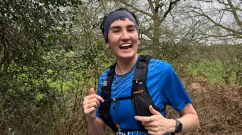 Alice Bolton The image shows a smiling woman running, wearing a blue top and black vest on top. There is a watch on her left wrist and trees can be seen behind her.