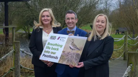 Ulster Wildlife Gina McIntyre, Andrew Muir and Dawn Miskelly pose for a launch photo at Lough Neagh Discovery Centre. McIntyre and Miskelly both have long, blonde hair and are wearing black suits. Muir has short, grey hair and is wearing a blue suit, blue shirt and a dickie bow. All three are holding a poster showing a photo of a bird. The poster says "Breathing life back into our peatlands". 