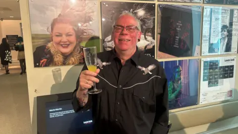 Jack Conlon/BBC A smiling man holding a drink in his hand which is raised in the air. He has grey hair and is wearing a black shirt and is stood in front of a wall with several pictures on.