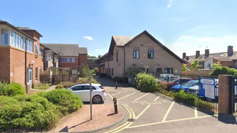 The former Pershore police station pictured from a car park. The building is fenced off by a metal gate and is made up of cobbled bricks. The roof has a triangle shape to it and the building is comprised of two floors. 