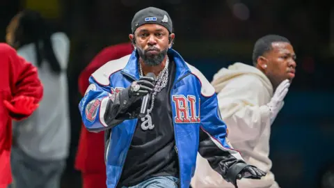 Gatety Images Raper-Centric Lammer 2025 Veste uma jaqueta azul do time do colégio e um boné de beisebol nas costas enquanto se apresenta na frente de coreógrafos no show do intervalo do Super Bowl.