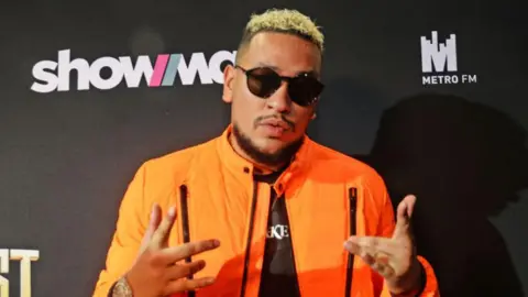 Gallo via Getty Images South African rapper Kiernan Forbes, popularly known as AKA, is seen in an orange jacket and glasses on while he attends an event