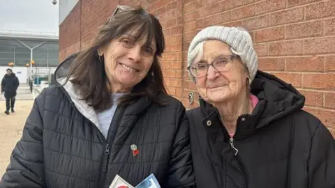 Joan Booth (left) has long brown hair and is wearing a grey sweat shirt and a long black black puffa coat. Joan Moffatt has white hair and dark spectacles and wears a black coat. They ae in front of a red brick wall across the road from large Tesco store.