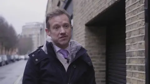 Ben Beadle is seen standing in the street next to a building's garage-like entrance. He is wearing a shirt and tie under a jumper and coat. A row of parked cars can be seen in the road behind him.