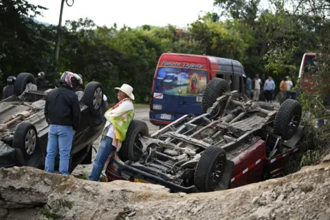 Jair Coll / Reuters Two people in a motorcycle helmet and white hat look on while surverying the scene which includes two upside down vehicles in front of a bus parked on the side of the road.