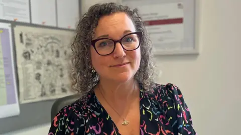 A woman sat in an office with shoulder-length brown hair and dark-rimmed glasses.